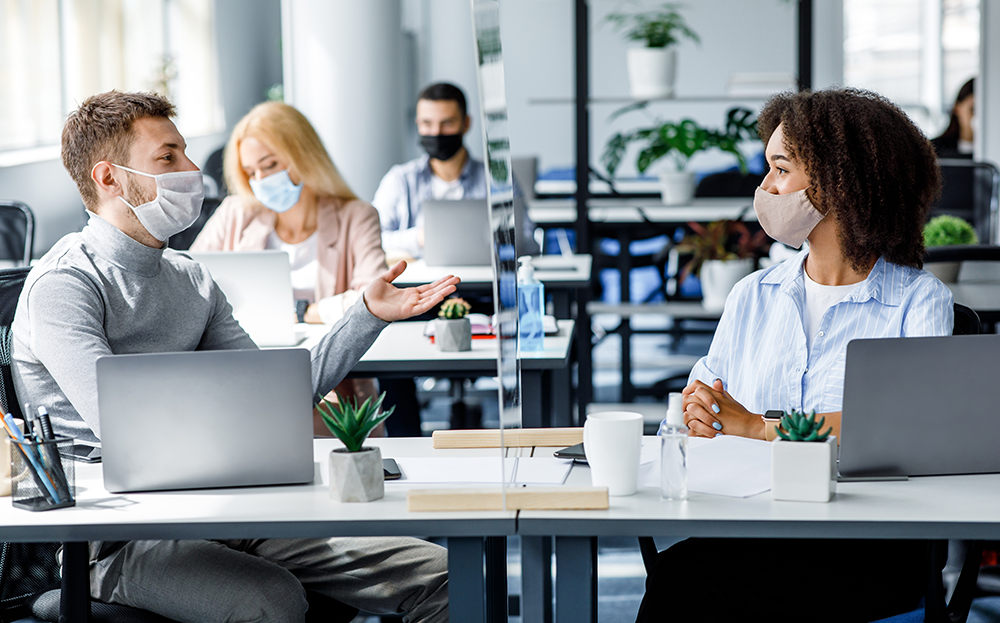 Two individuals wearing masks and conversing at the office with open laptops