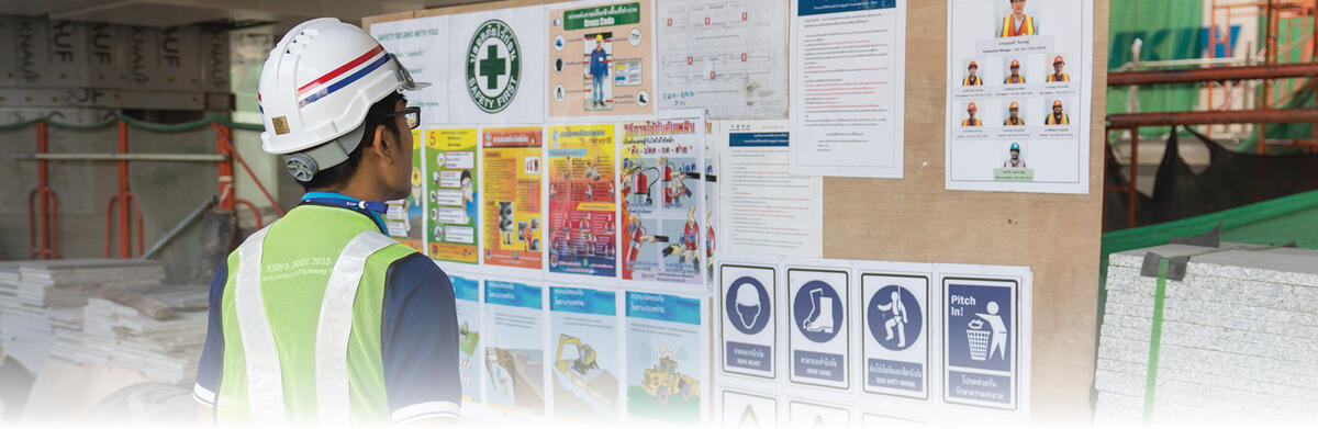 Worker in a yellow vest with a white hard hard looking at a bulletin board of safety announcements 