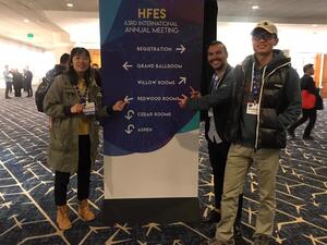 3 Students pointing a banner at the HFES 2019 conference