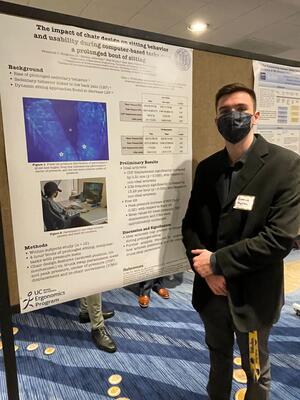 Freddie Houghton standing next to his poster on "The impact of chair design on sitting behavior and usability during computer-based tasks over a prolonged bout of sitting"